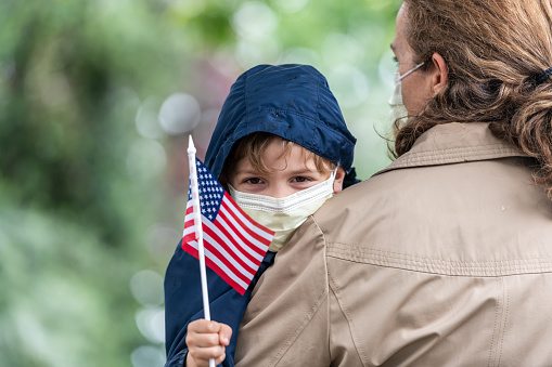 Mature woman carrying her son, both wearing protection masks, the child is holding a US flag